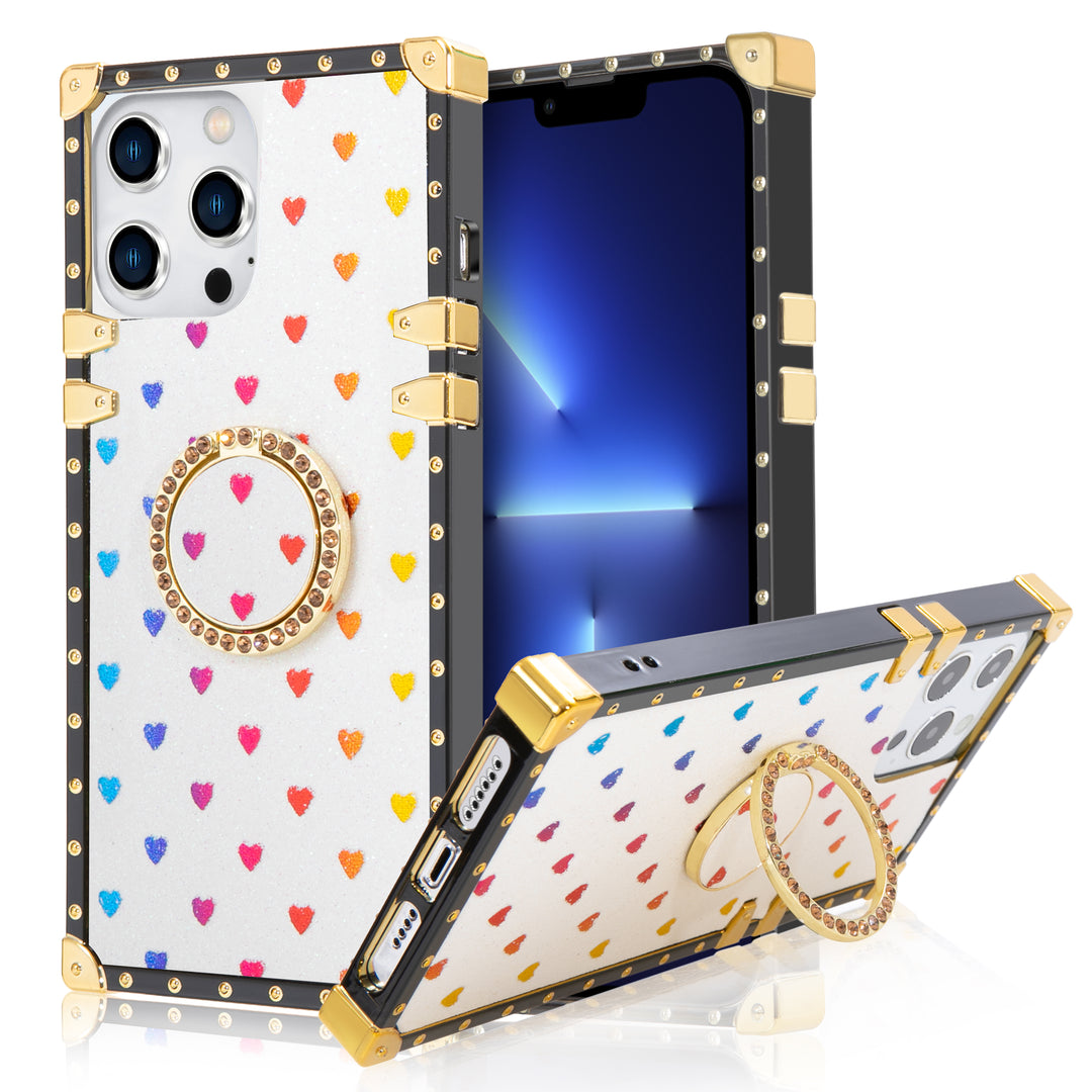 A box-shaped, TPU, iPhone 12 Pro Max case, with gold corner bumpers and gold rivets on its black trim. The case’s back is designed white, with rainbow hearts, and glitter. A gold trim ring, with diamond studs, doubles as a handle and kickstand for the case. #color_white-glittery-hearts